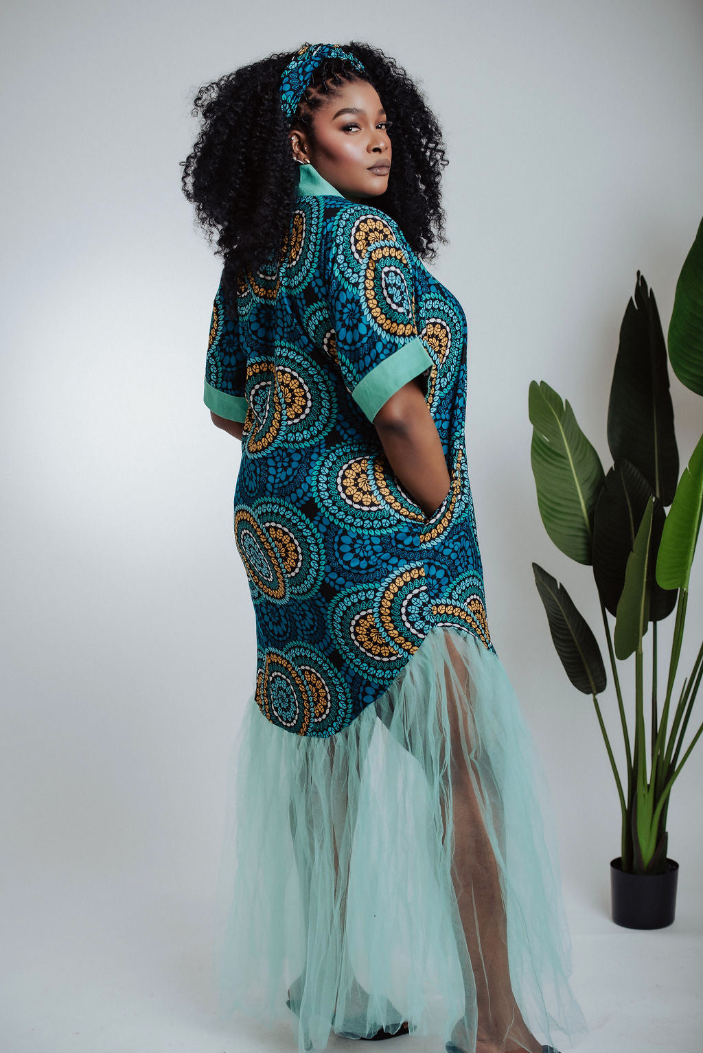 Woman wearing teal traditional African print dress/boubou with a geometric pattern and tulle with a plant in the background - back view