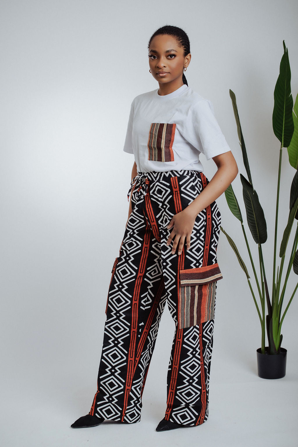 Woman wearing a white t-shirt with a patterned pocket and geometric African print cargo pants standing next to a plant.