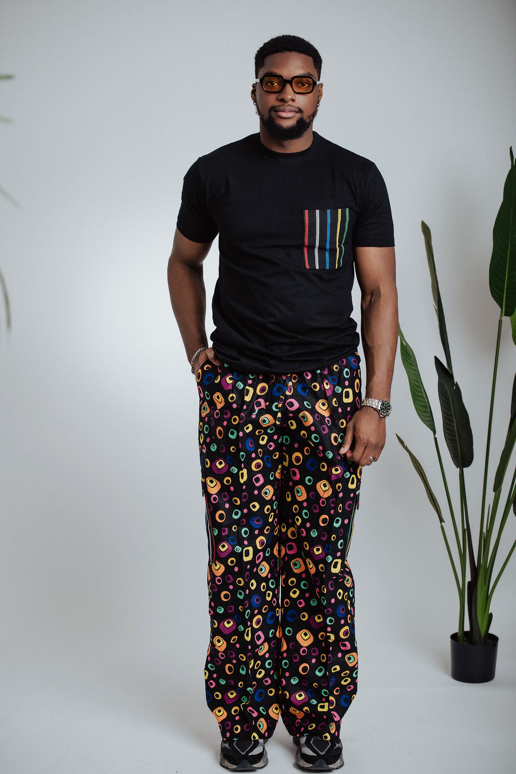 Man wearing a black t-shirt with colorful pocket design and patterned cargo pants standing against a white background with a plant