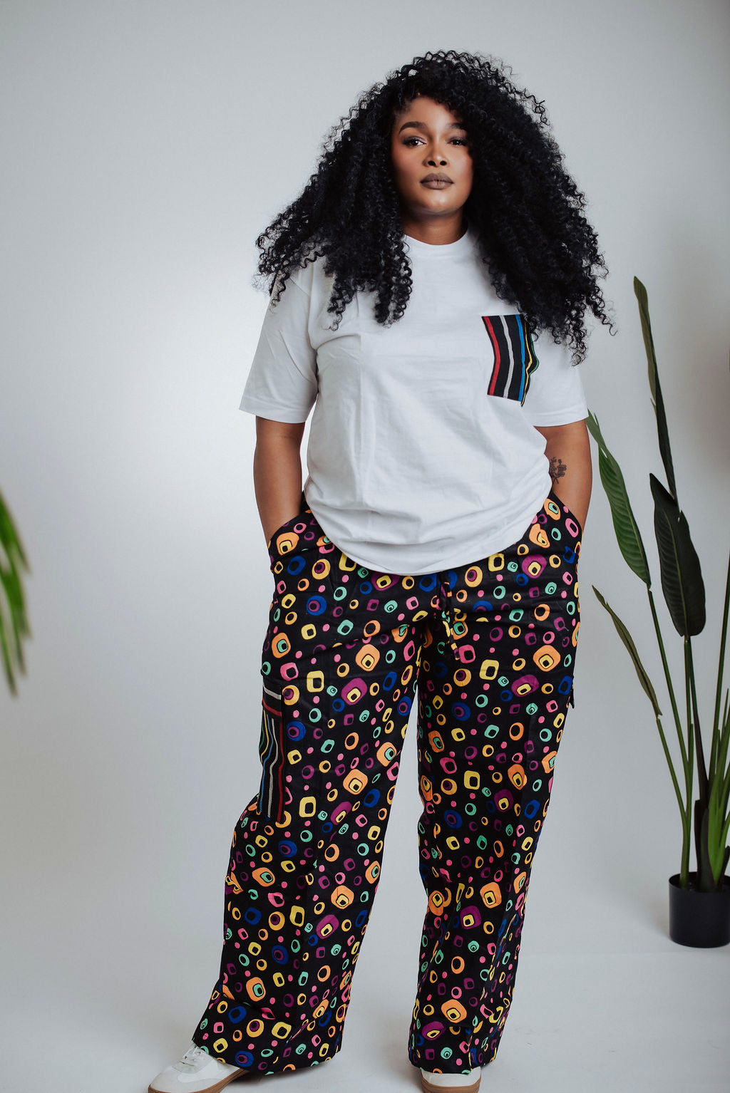 Female model wearing a white t-shirt with a colorful pocket design and black cargo pants with a multicolor pattern, standing against a plain background.