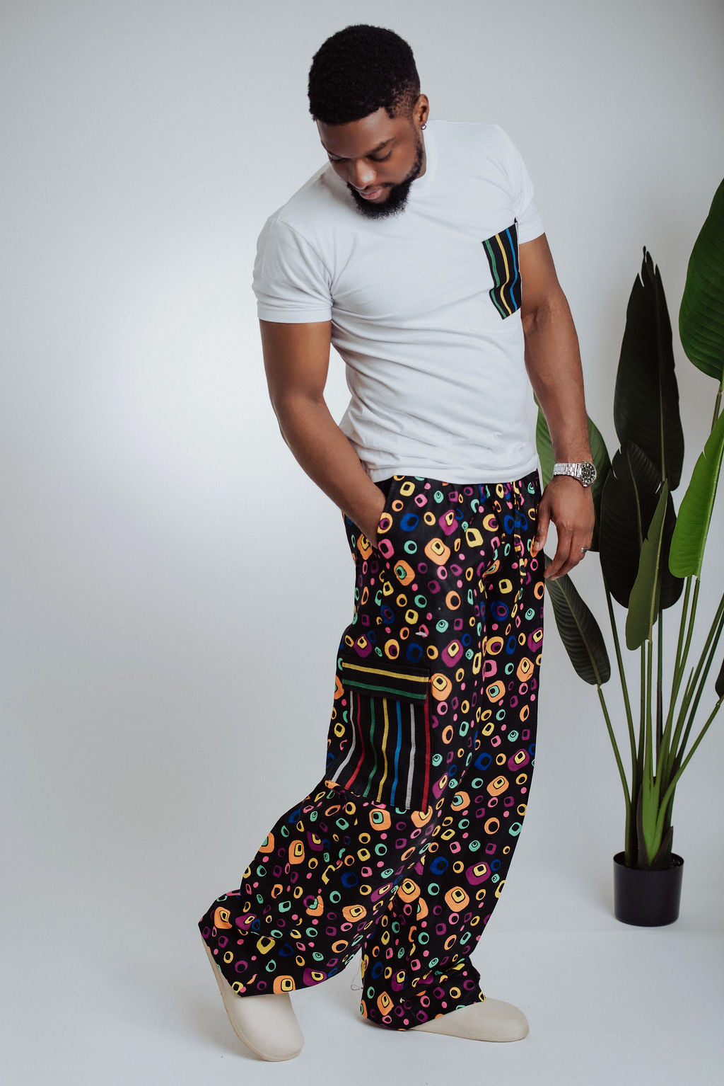 Man wearing a white t-shirt and colorful patterned African print cargo pants standing against a plain background with a plant.