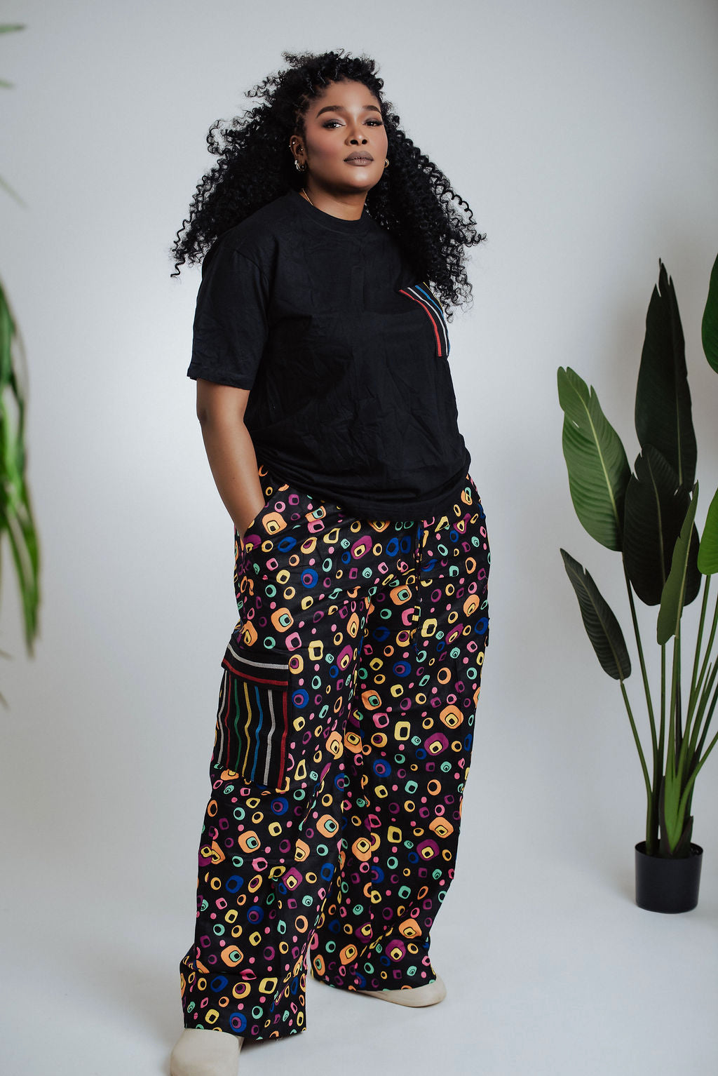 Female model wearing a black t-shirt and colorful African print patterned cargo pants standing against a white background with plants.