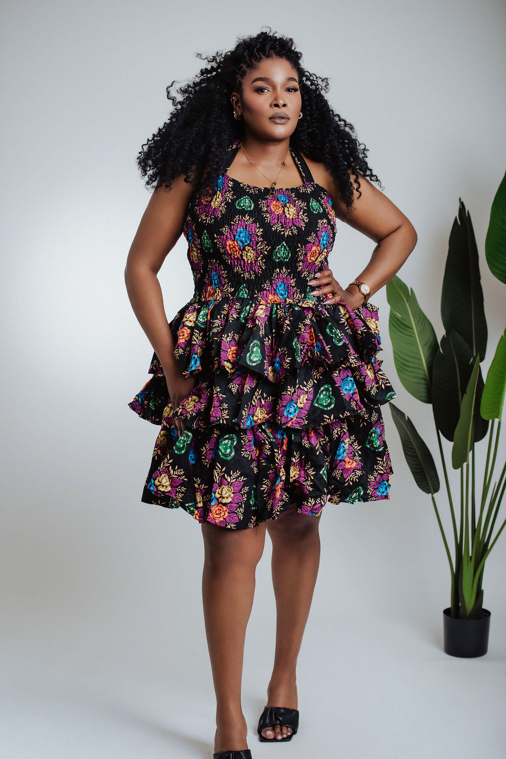 Woman wearing a colorful floral dress standing against a white background with a plant to the side.