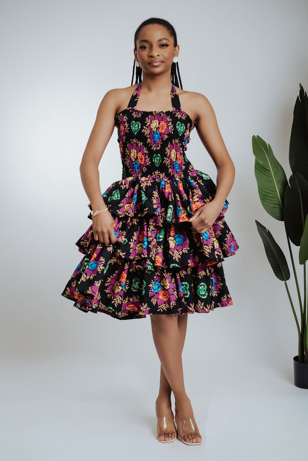 Woman wearing a colorful floral dress standing against a white background with a plant to the side.