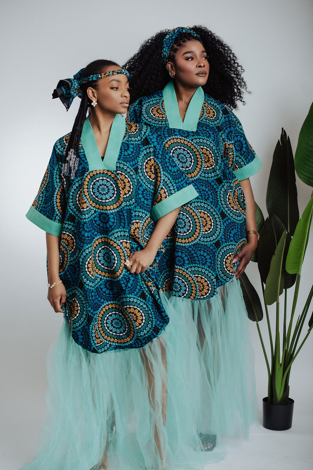 Two women wearing teal traditional African print dress/boubou with a geometric pattern and tulle on a plain background