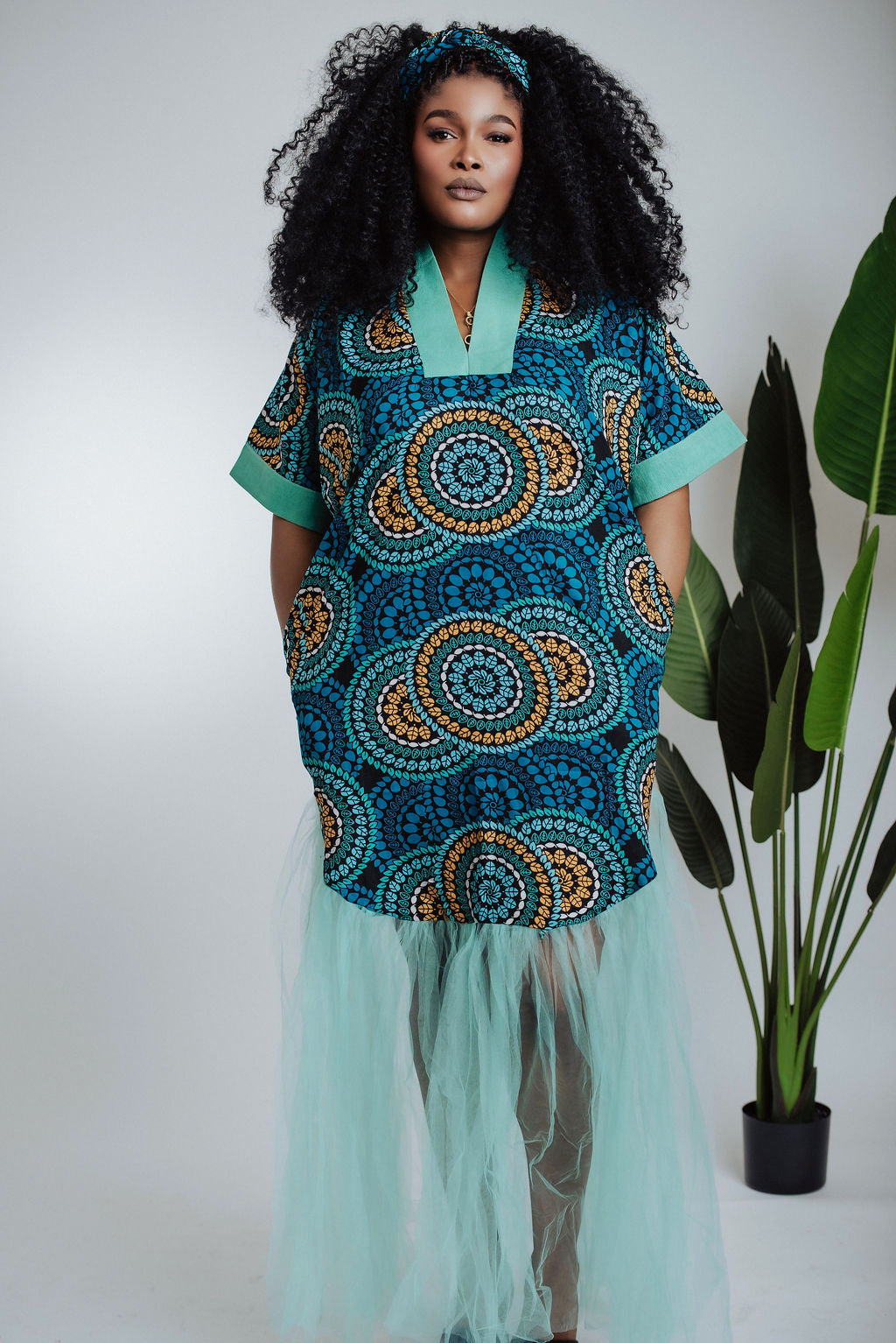 Woman wearing teal traditional African print dress/boubou with a geometric pattern and tulle with a plant in the background