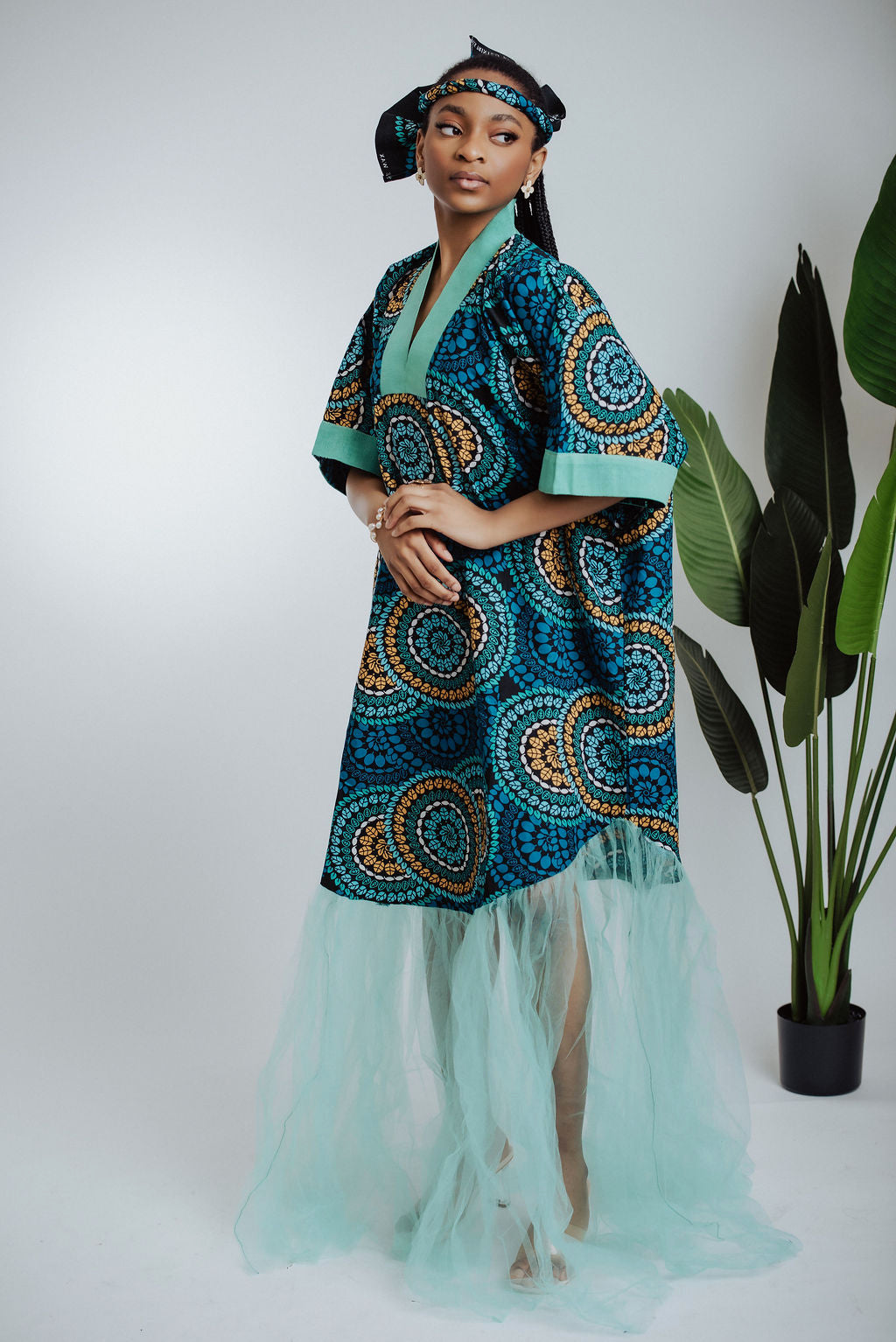 Woman wearing teal traditional African print dress/boubou with a geometric pattern and tulle with a plant in the background - side view
