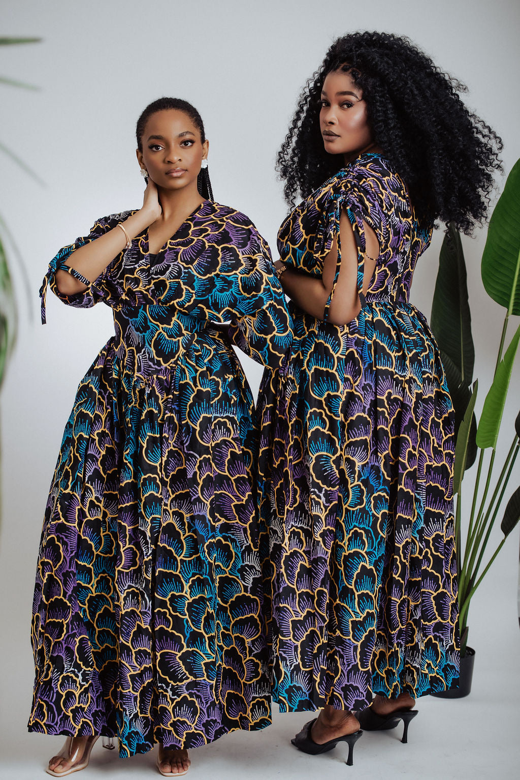 Two women wearing matching African print patterned maxi dresses standing side by side against a white background with plants.