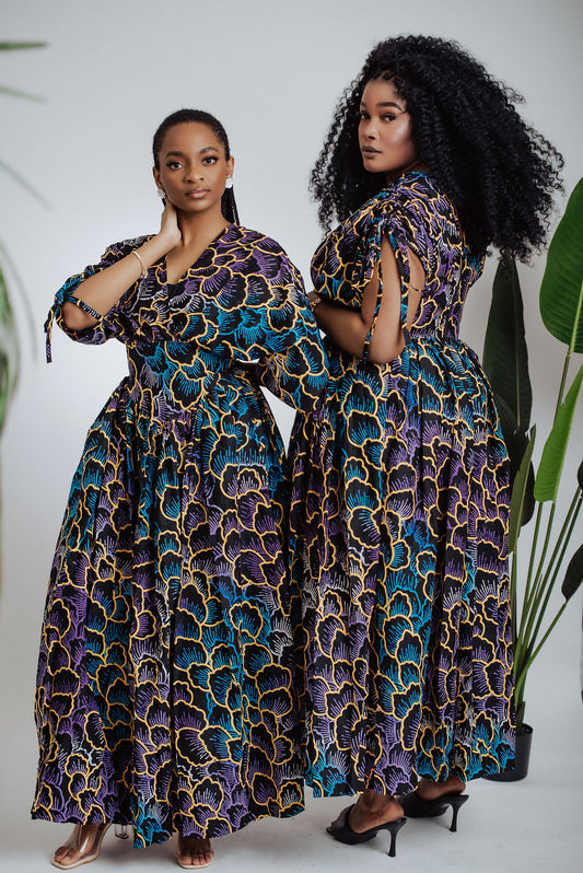 Two women wearing matching African print patterned maxi dresses standing side by side against a white background with plants.