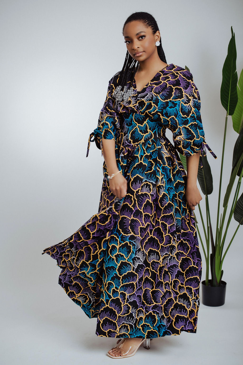 Woman wearing a long, patterned African print dress standing next to a plant on a white background