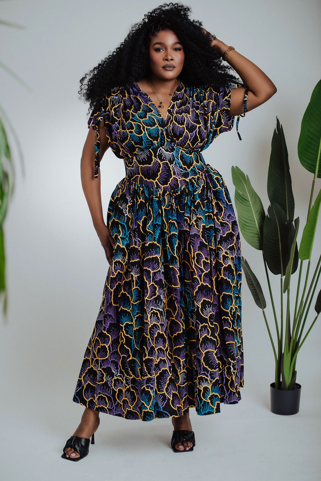 Woman wearing a patterned African print  dress standing next to a plant on a white background