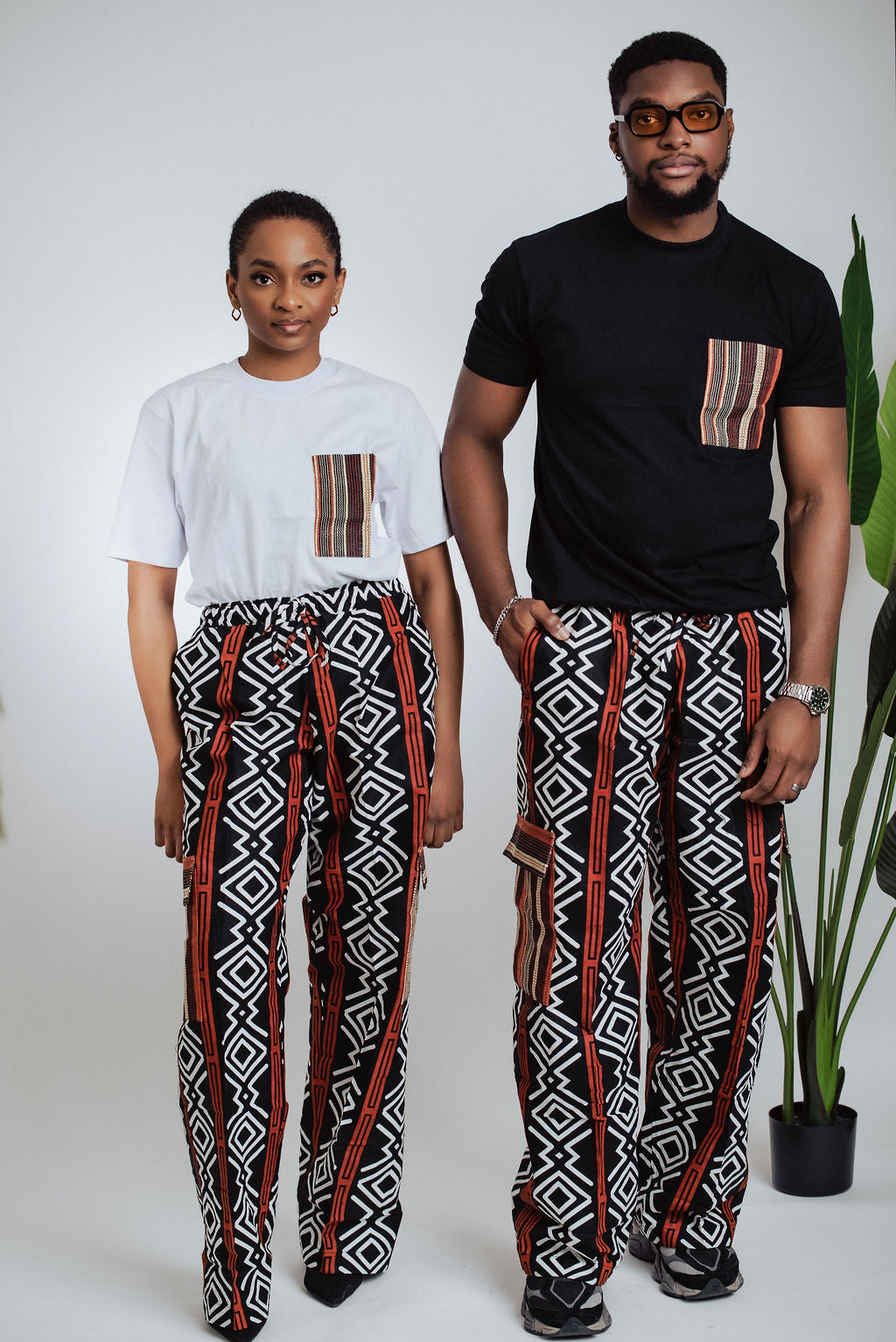 Male and female models wearing geometric African print cargo pants with black and white pocket T-shirts against a white background with a plant