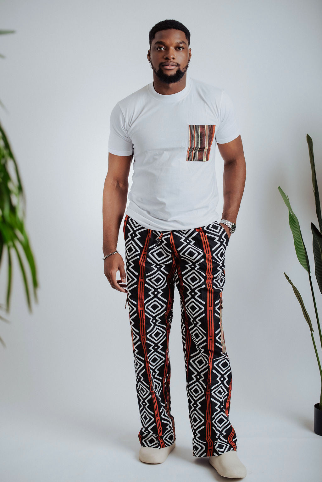 Man wearing a white t-shirt with tribal pocket design and geometric cargo pants standing against a white background with plants