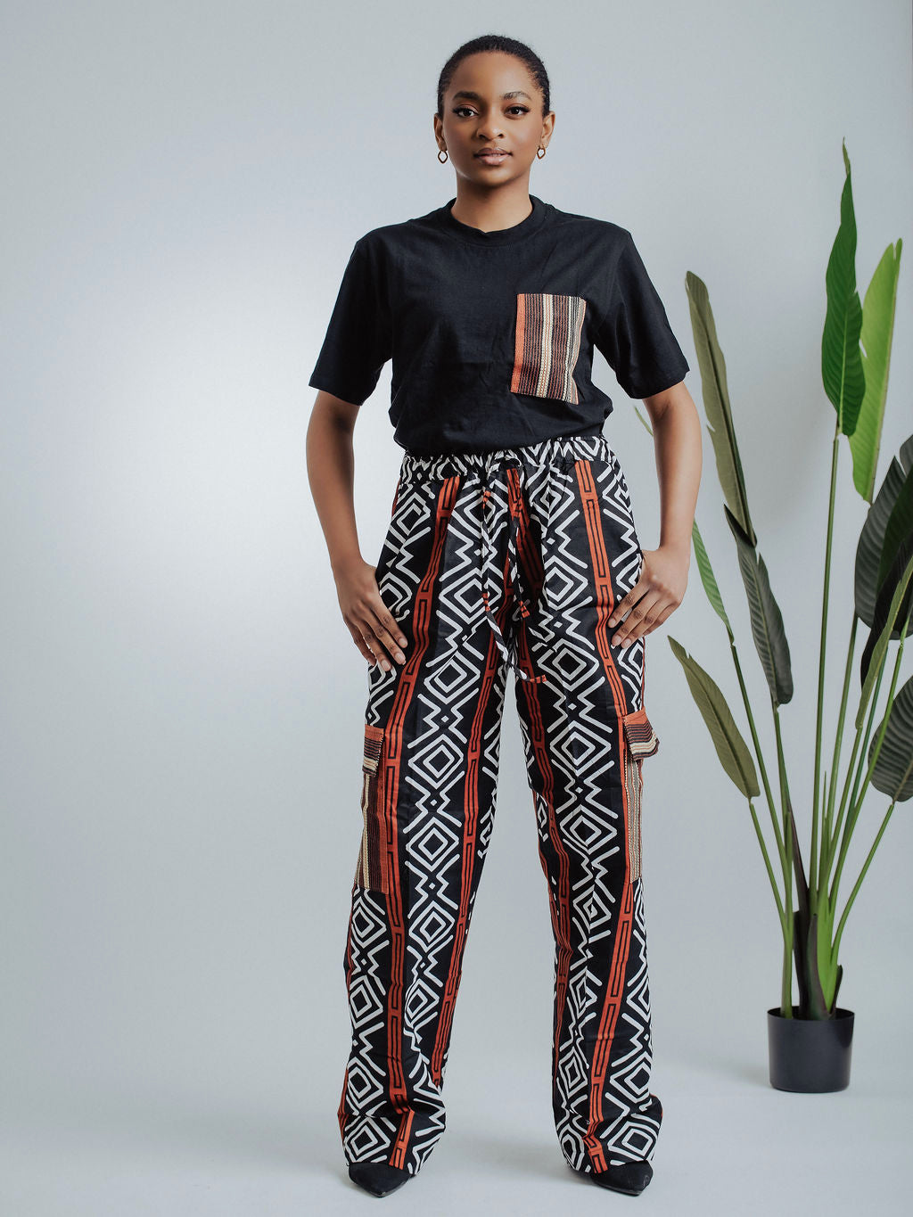 Female model wearing a black t-shirt with tribal pocket design and geometric cargo pants standing against a white background with a plant