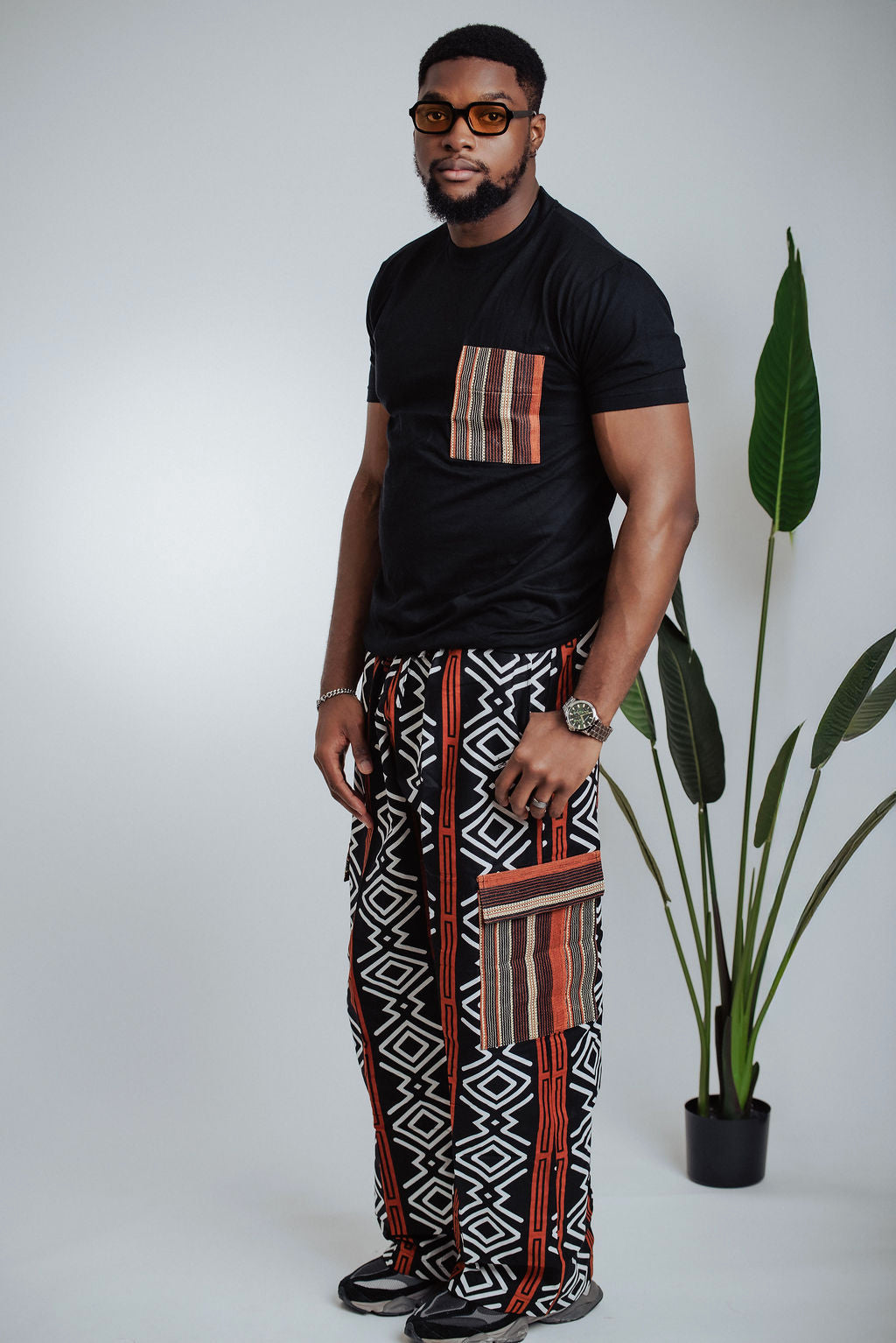Man wearing a black t-shirt with a patterned pocket and geometric African  print cargo pants standing next to a plant.