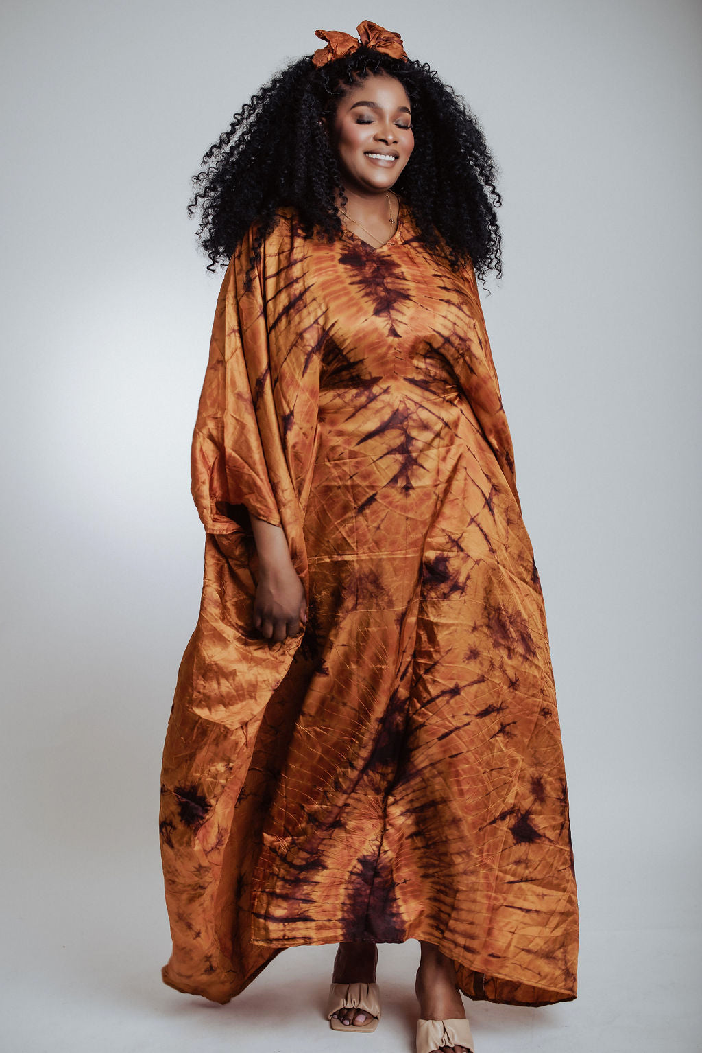 Woman wearing a long, silk tie-dye brown dress against a plain background